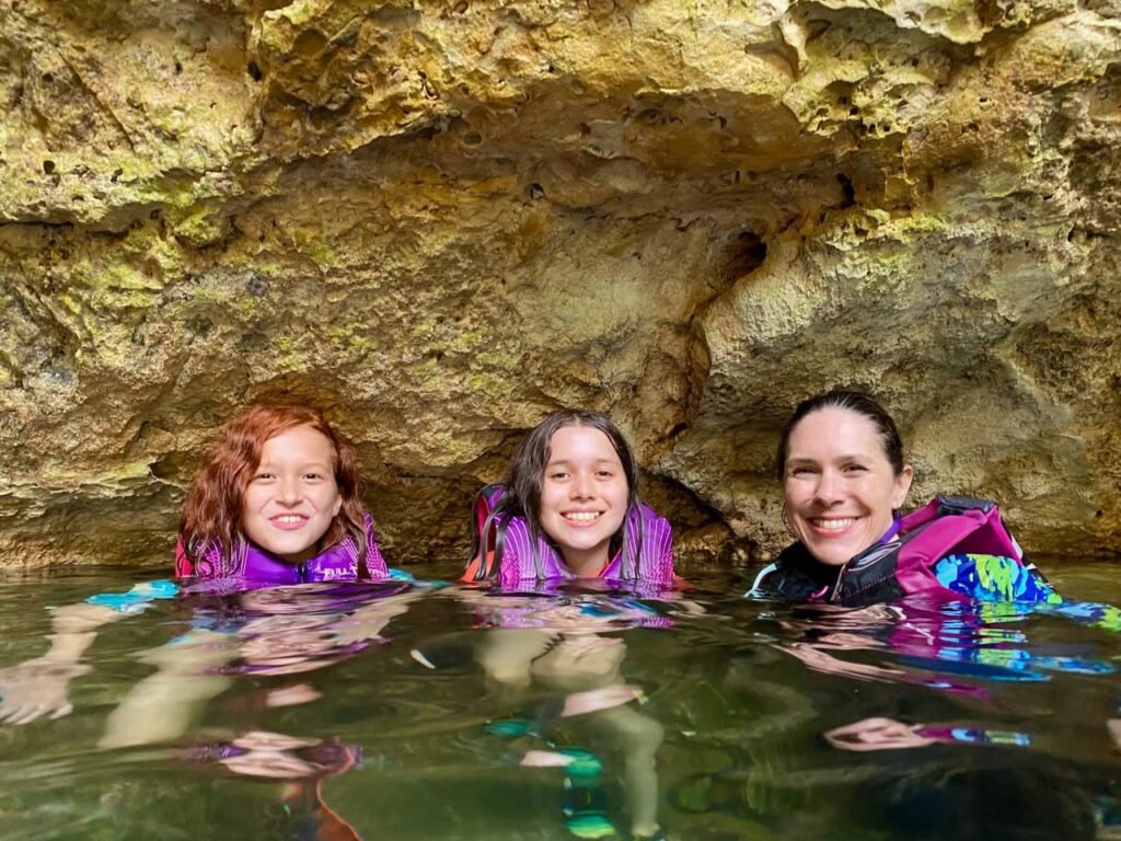 A group of three people wearing life jackets floats in calm, shallow water inside a rocky cave. The image pairs a wide shot with close-up details of the stone formations and people.