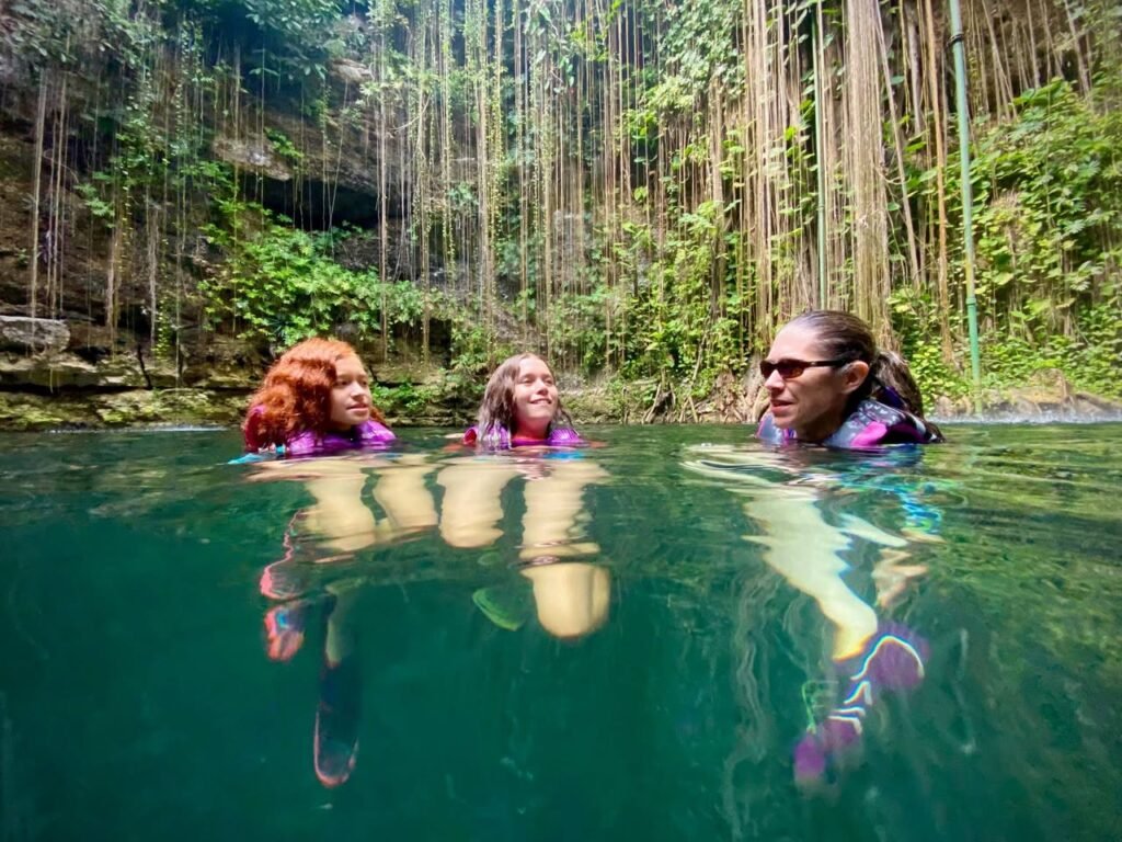 Travellers swim in a clear blue-green cenote surrounded by lush jungle vegetation.
