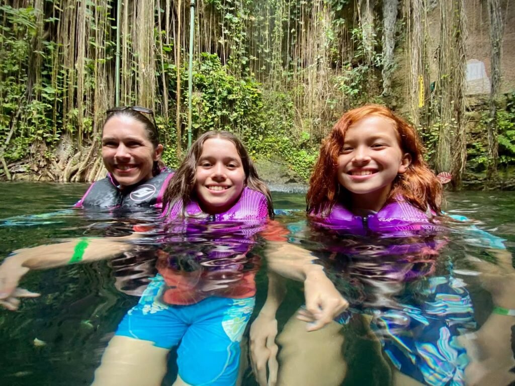 Three people swim smiling in a clear jungle river, wearing life vests with green vines in the background.