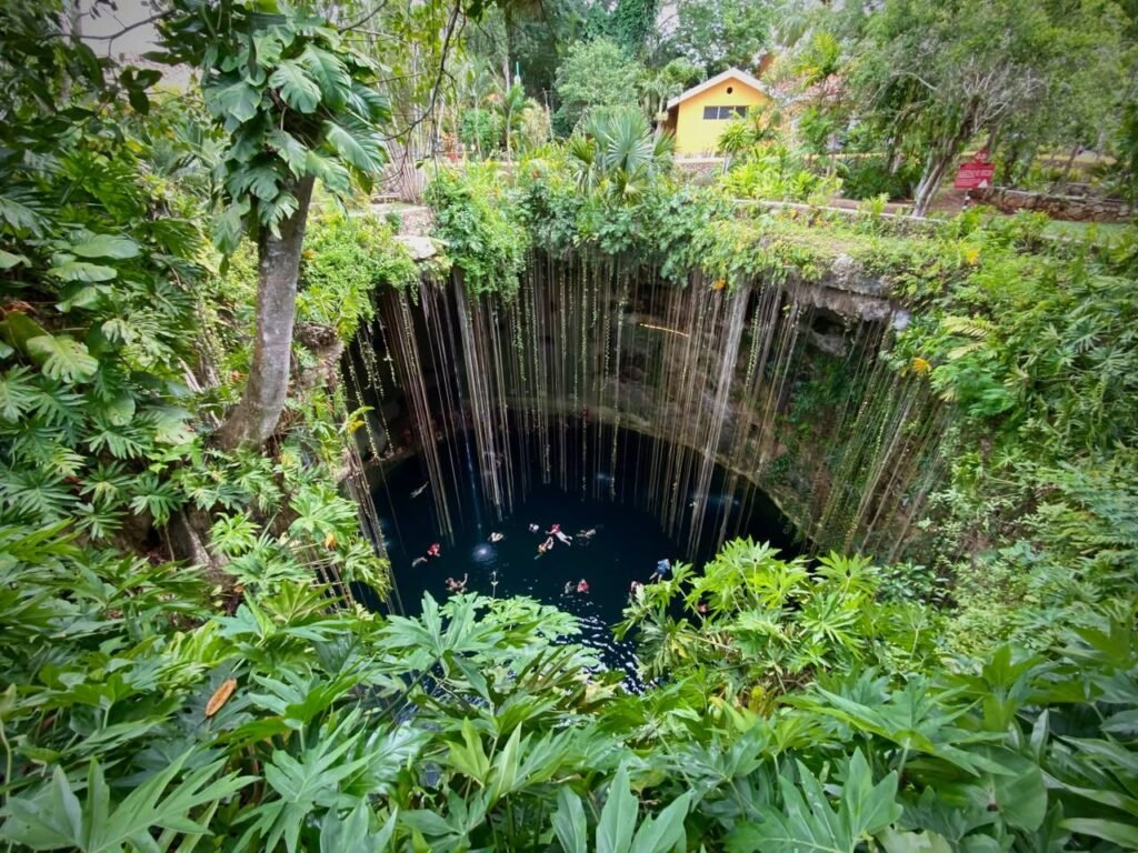 A deep blue cenote surrounded by dense green foliage with a yellow building perched along the rocky rim.