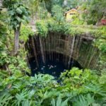 Coba Ruinas Arqueológicas A deep blue cenote surrounded by dense green foliage with a yellow building perched along the rocky rim.