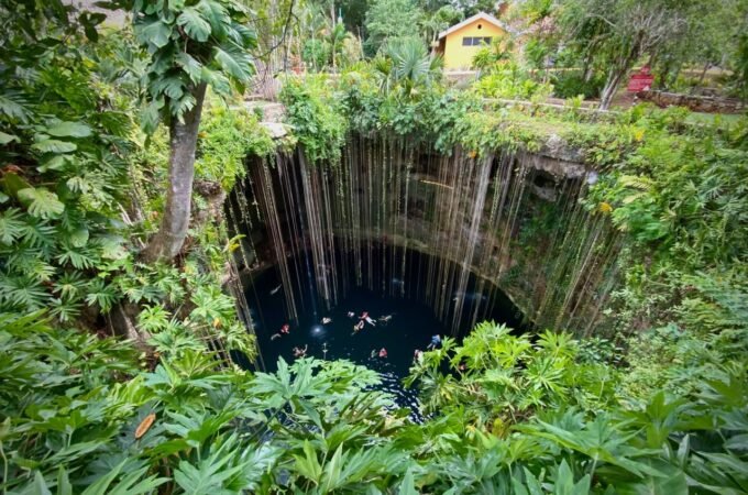 A deep blue cenote surrounded by dense green foliage with a yellow building perched along the rocky rim.