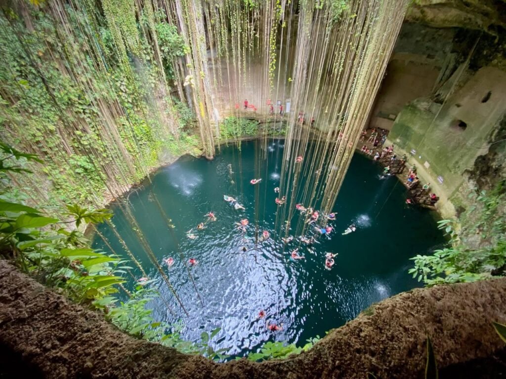 A stunning view of Cenote XCANAH with swimmers enjoying the deep blue water as vines cascade down the lush walls.