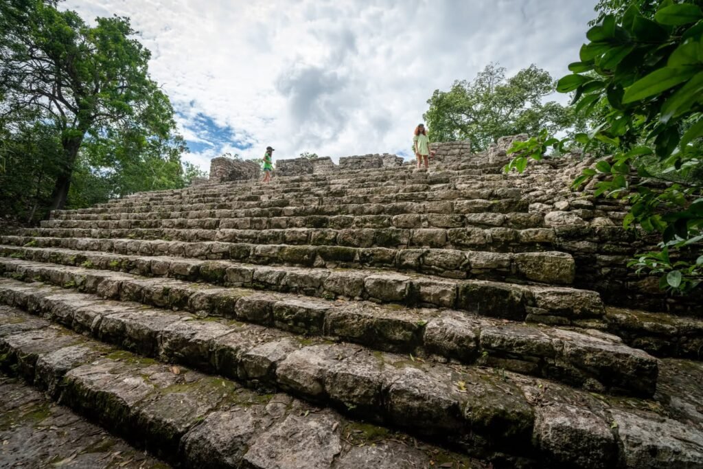 A low-angle view of weathered stone steps on a large mayan pyramid. Lush green trees frame the sides against a cloudy sky.