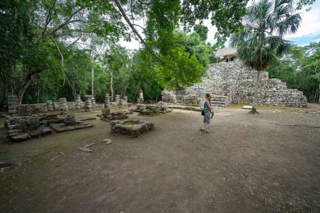 A photo of ancient stone ruins surrounded by dense tropical forest. A person stands near the structures in the background.