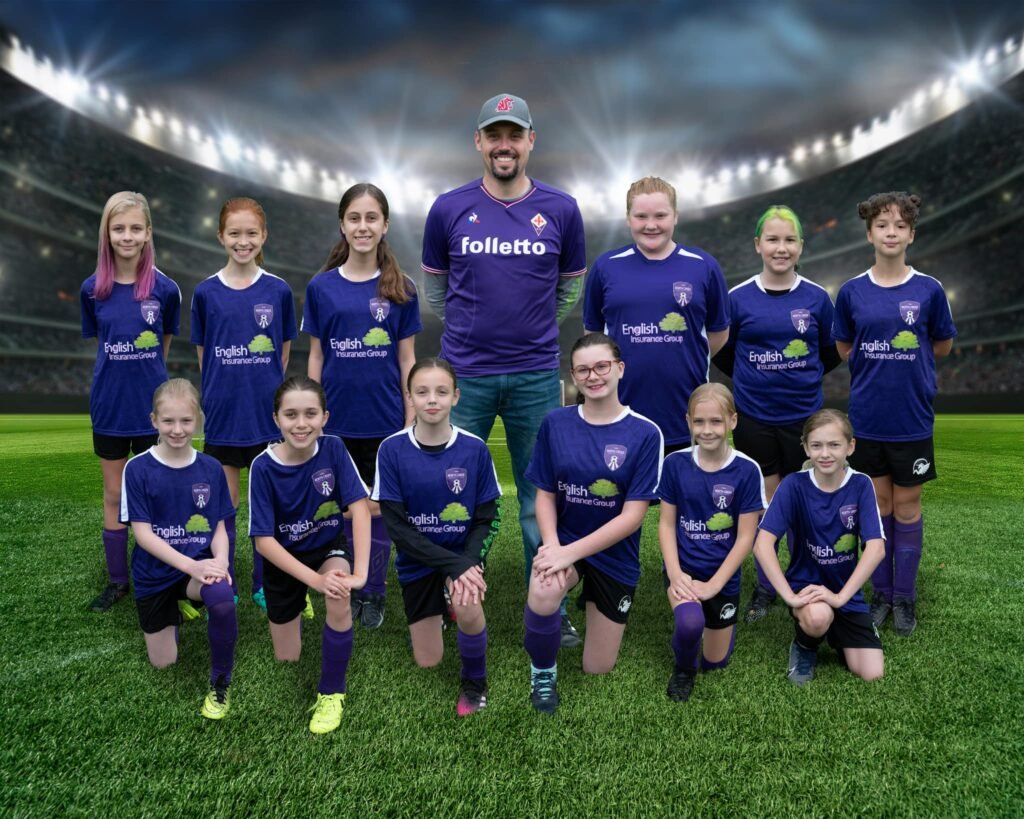 A youth soccer team in purple uniforms poses for a group photo on a grassy field with stadium lights in the background. A team photo in purple soccer uniforms at a stadium backdrop.