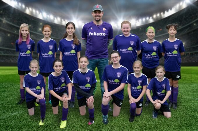 A youth soccer team in purple uniforms poses for a group photo on a grassy field with stadium lights in the background. A team photo in purple soccer uniforms at a stadium backdrop.