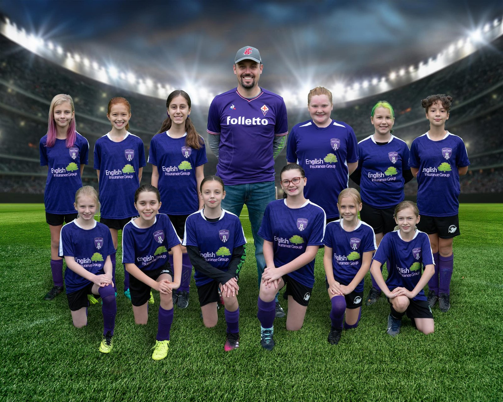 A youth soccer team in purple uniforms poses for a group photo on a grassy field with stadium lights in the background. A team photo in purple soccer uniforms at a stadium backdrop.