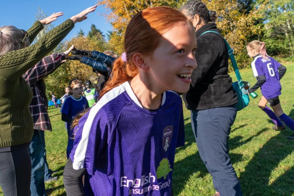 A group of girls in purple soccer uniforms and spectators gather on a sunny field. One girl smiles as others raise their arms in celebration.
