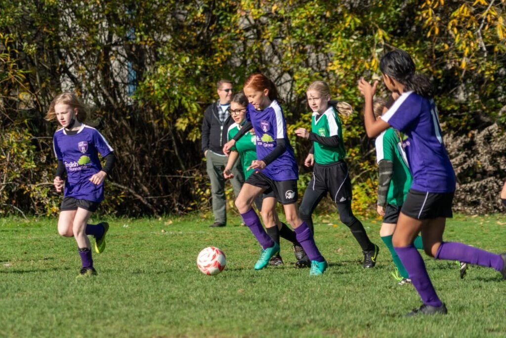 Youth soccer players in purple and green uniforms compete during a match on a grassy field surrounded by autumn foliage.