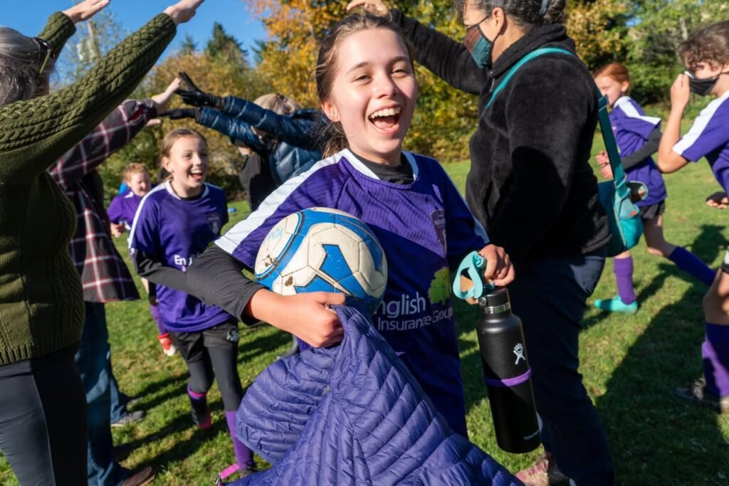 A young soccer player in a purple uniform holding a soccer ball with teammates and adults celebrating outdoors.