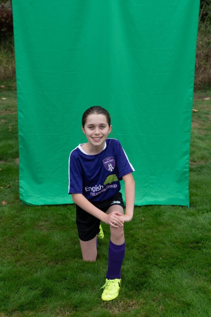 A person wearing a purple soccer uniform kneeling on a grassy field in front of a bright green screen, preparing for a photo session