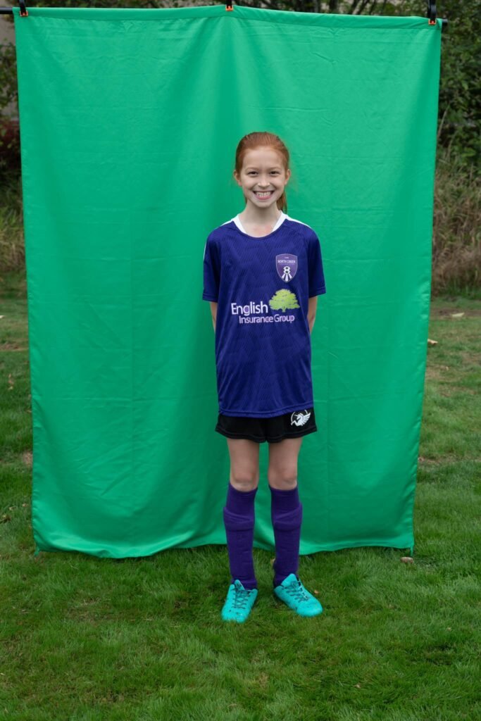A young soccer player wearing a purple uniform and turquoise cleats posing in front of a green backdrop on a grassy field.