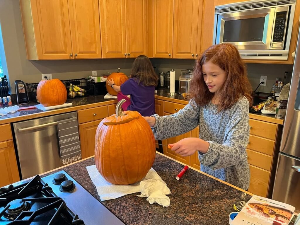 A child carving a pumpkin in a wood-paneled kitchen. Two people are working with pumpkins on a granite countertop near stainless steel appliances.