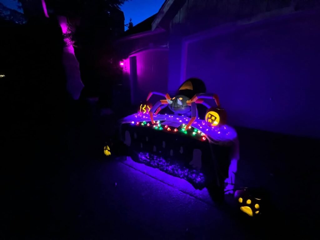 A brightly lit Halloween decoration glowing in purple and green on a dark street at night.