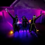 Floating pumpkin-shaped drones with lit interiors glowing in purple light. People with neon face paint dance among floating pumpkin lights.