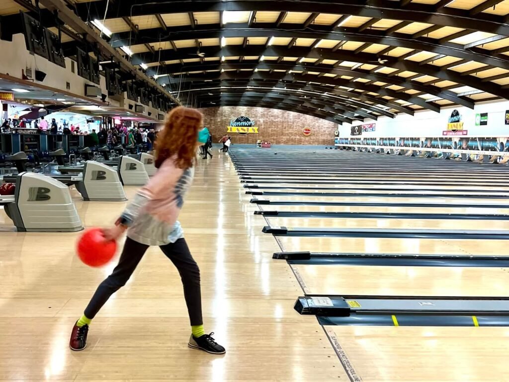 A person in a dynamic bowling stance at a crowded bowling alley with a red ball in hand.