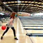 Drogon A person in a dynamic bowling stance at a crowded bowling alley with a red ball in hand.
