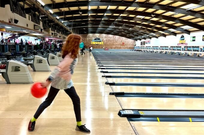 A person in a dynamic bowling stance at a crowded bowling alley with a red ball in hand.