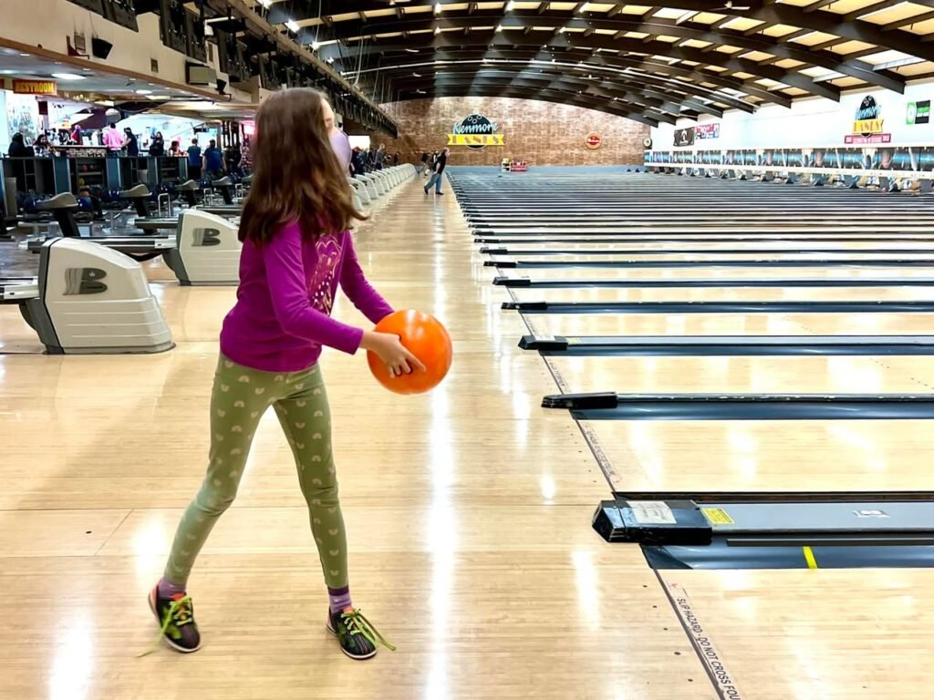 A person in colorful pants holds an orange bowling ball at an indoor bowling alley. The background shows multiple lanes and bustling activity in the distance.