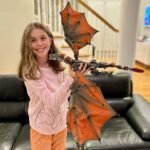 A girl in a living room smiles while holding a large robotic dragon drone with vibrant orange wings.