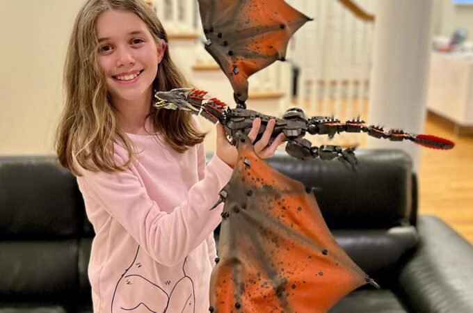 A girl in a living room smiles while holding a large robotic dragon drone with vibrant orange wings.