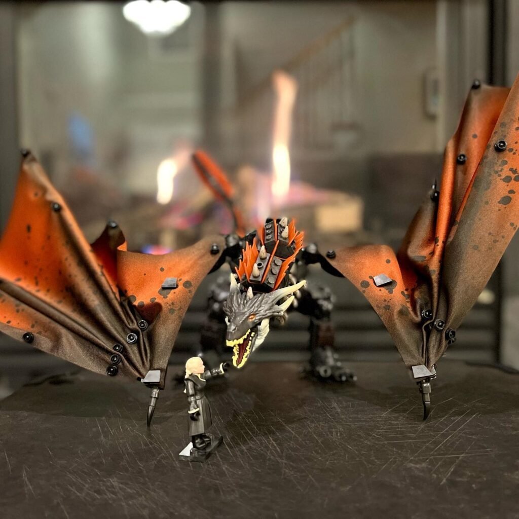 A detailed model of a mechanical dragon with orange wings overlooking a human figure. A second view shows a printing or assembly process with the dragon's wing in the foreground.