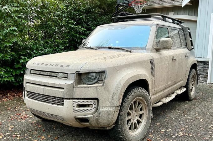 A mud-covered Land Rover Defender parked in a driveway. The vehicle shows signs of recent off-road adventure in the Pacific Northwest.