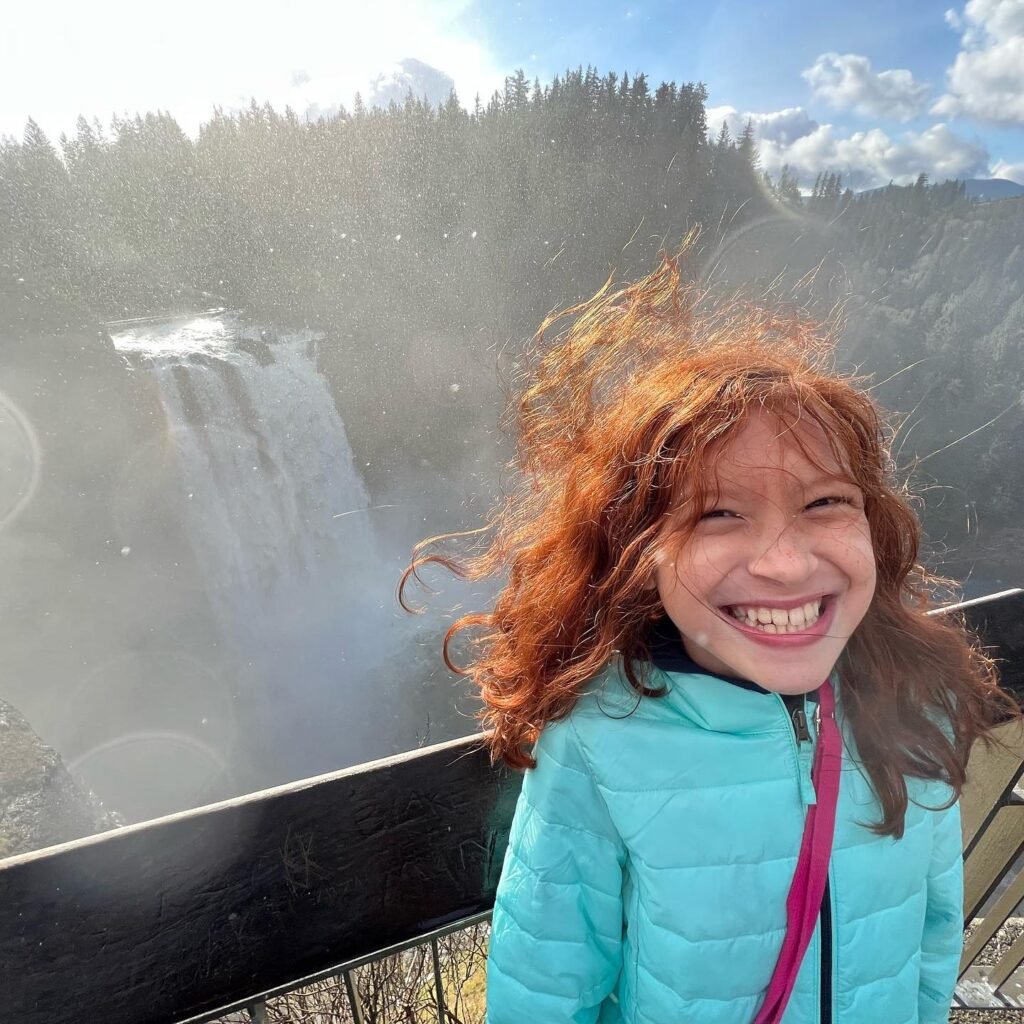 A person with red hair smiles broadly in a light wind at Multnomah Falls in the Pacific Northwest. The misty waterfall and forested hills create a scenic background.
