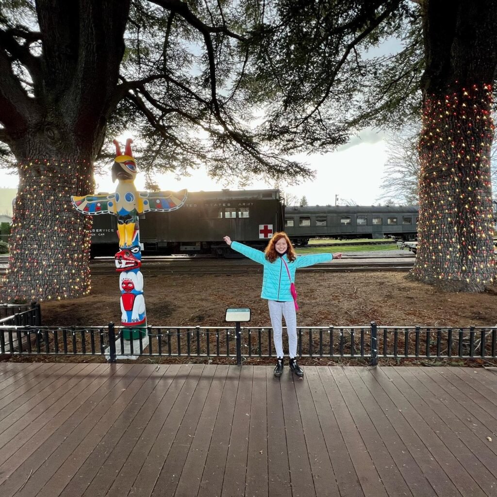 A person spreads their arms joyfully on a wooden deck at a railway museum, with vintage train cars and illuminated totem poles in the background.