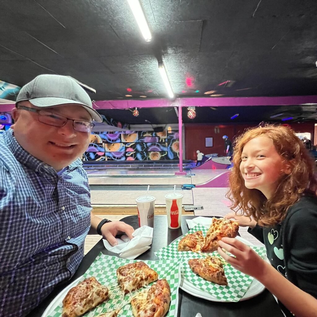 Two people enjoy pizza and drinks at a vibrant bowling alley with colorful graffiti art.