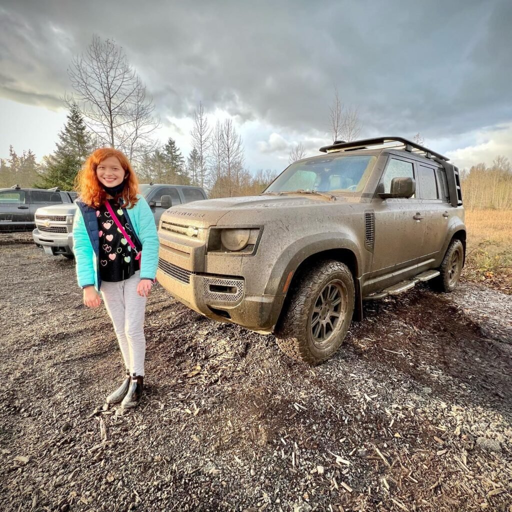 A person poses beside a muddy Land Rover in a Pacific Northwest forest setting. The vehicle is covered in mud, suggesting an off-road adventure in the wild.