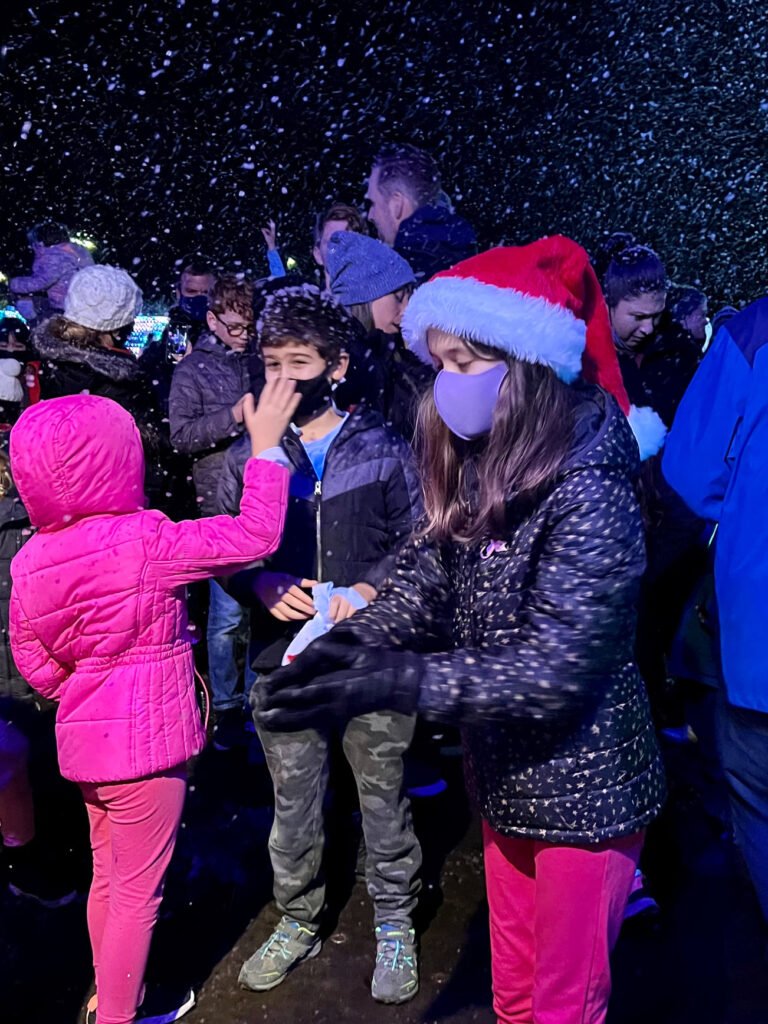 Festival-goers bundled in colorful winter coats gather under artificial snow during an outdoor holiday event. One person wears a festive Santa hat while enjoying the atmosphere.