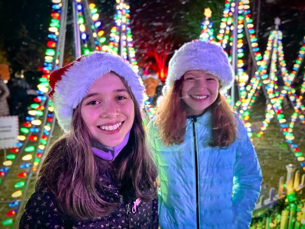 Two smiling children wearing winter hats stand in front of colorful light displays on illuminated stilt structures. They appear to be at a festive seasonal event.