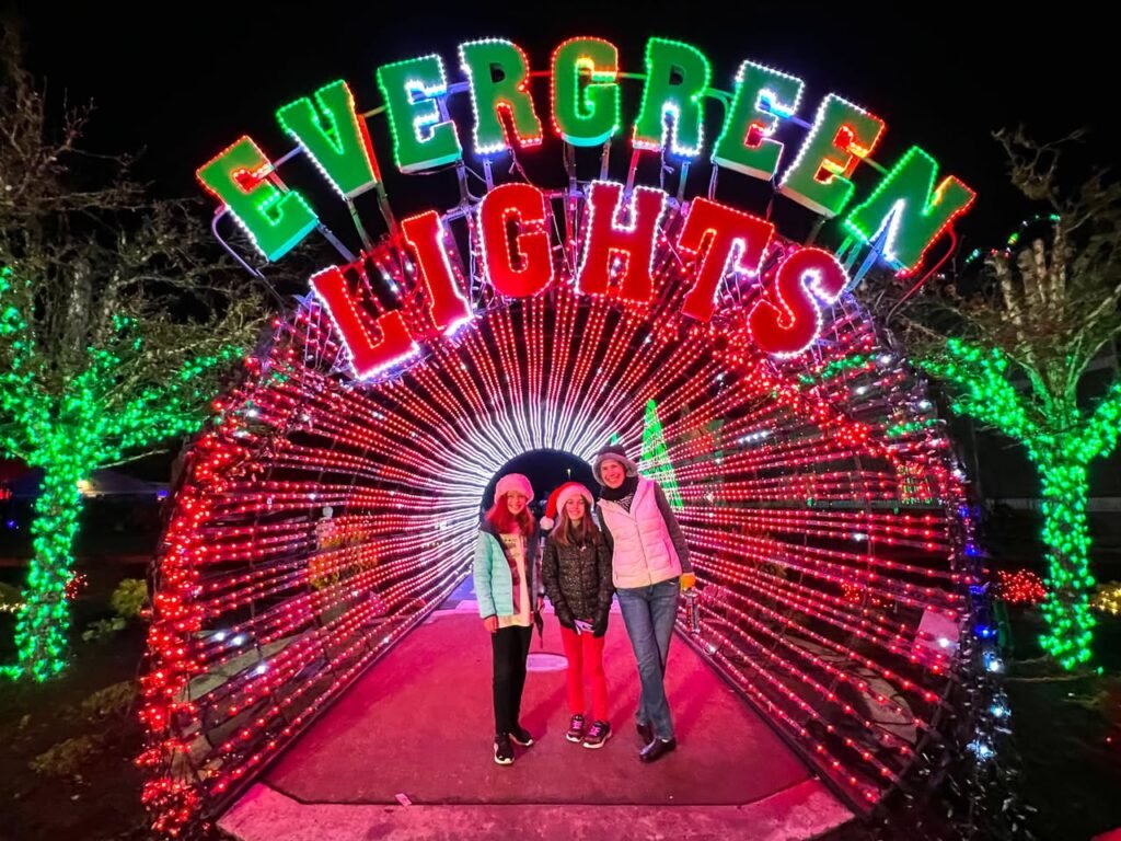A group poses in a tunnel of red lights under the colorful illuminated sign at Evergreen Lights. Green tree lights frame the festive holiday scene at night.