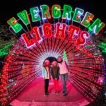 Suncadia Christmas 2021 A group poses in a tunnel of red lights under the colorful illuminated sign at Evergreen Lights. Green tree lights frame the festive holiday scene at night.