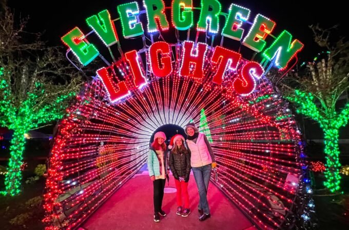 A group poses in a tunnel of red lights under the colorful illuminated sign at Evergreen Lights. Green tree lights frame the festive holiday scene at night.