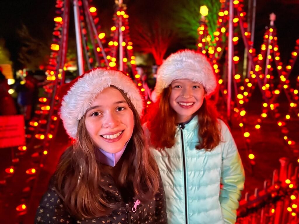 Two people wearing white fluffy hats and winter coats stand together with festive holiday lights in the background.