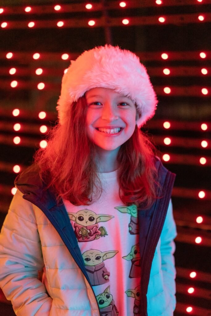 A smiling person wearing a fluffy hat and a light-up top in front of red lights. The background features rows of glowing red bulbs creating a festive mood.