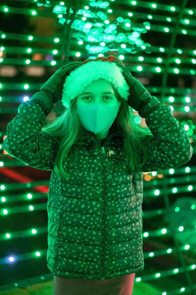 A person in a winter hat and coat stands before a wall of bright green holiday lights.