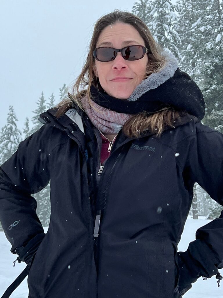 A person posing in winter gear amidst a snowy landscape with evergreen trees. Snowflakes are falling gently around them.