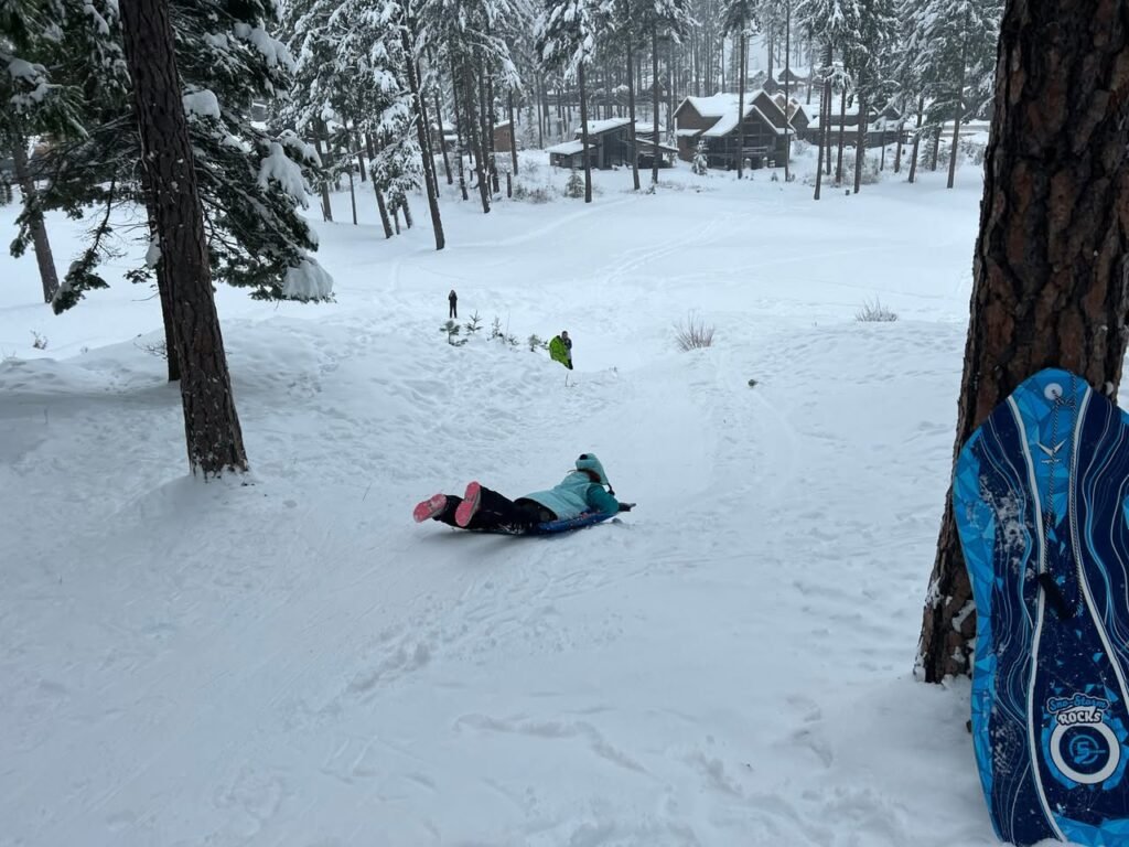 A person lies prone on the snow with a blue snowboard leaning against a pine tree, with cabins visible in the background.