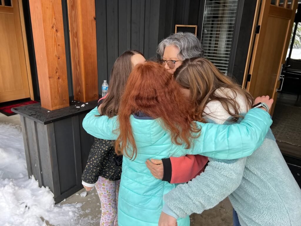 A family shares a warm group hug on a porch with snow. The setting includes wooden siding and an outdoor door.