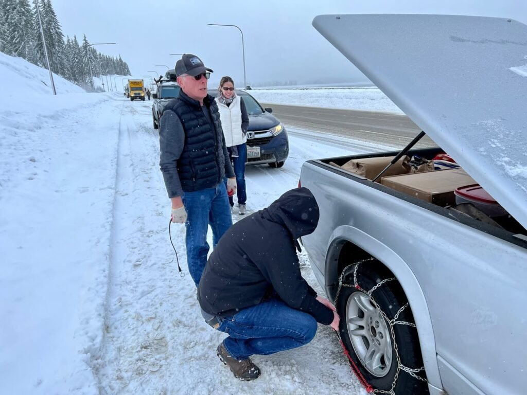 A person crouching to examine a car tire with chains on a snowy road. In the background, others stand near parked vehicles on a snow-covered highway beside the Pacific Northwest shoreline.