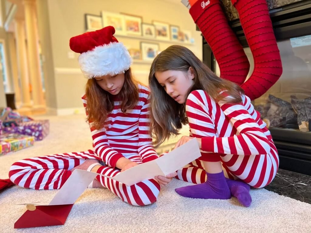 Two young girls in red and white stripping pajamas and a santa hat sit on the floor by the fireplace.
