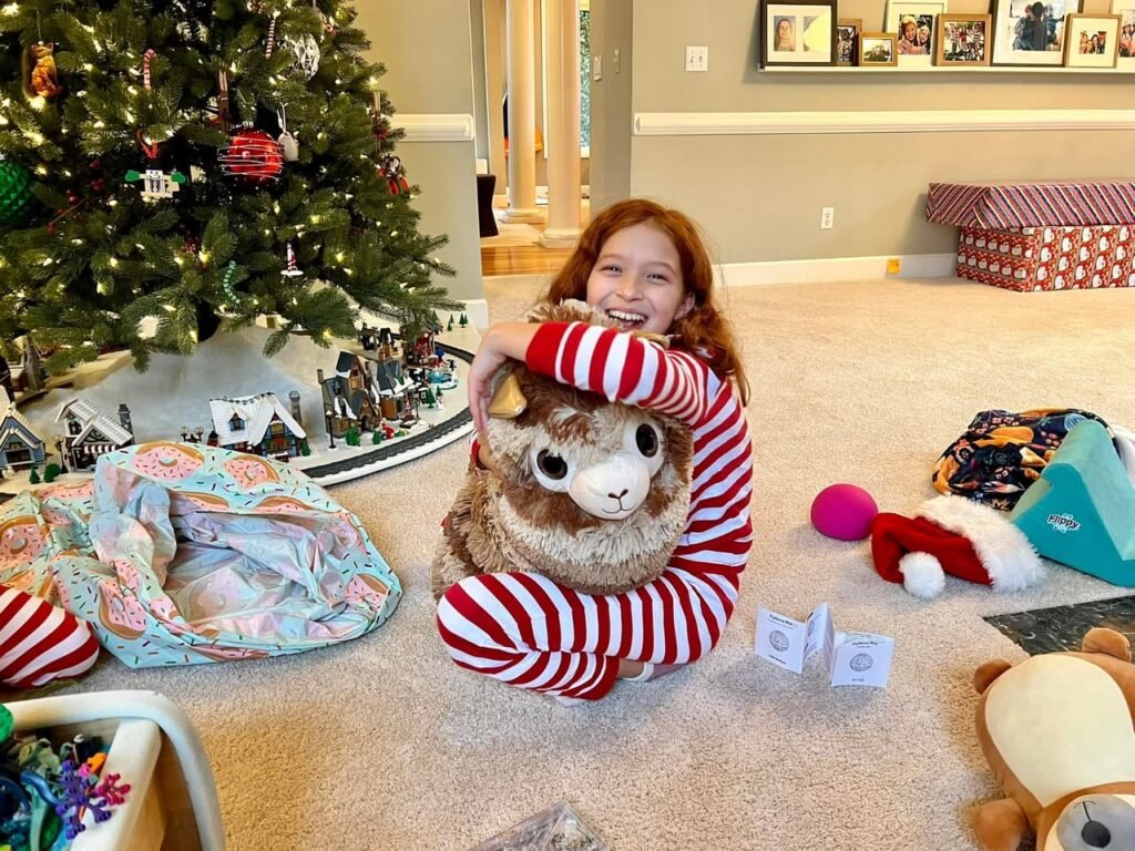 A child holds a large stuffed animal in front of a decorated Christmas tree in a home setting.
