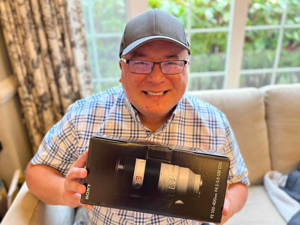 A smiling person holds a new Sony lens box indoors. The scene includes a beige sofa and windows with natural light.