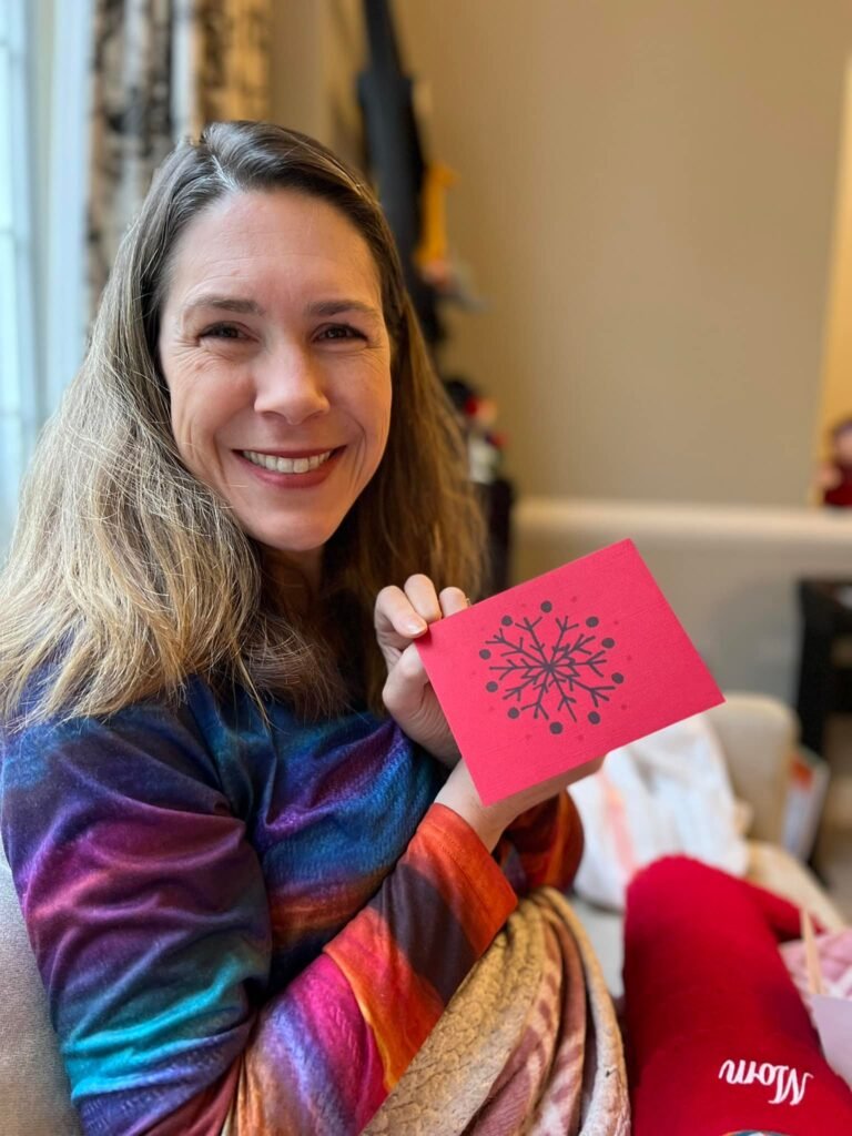 A woman smiles warmly while holding a pink card with a black snowflake design. She wraps herself in a vibrant, multicolored blanket in a home setting.