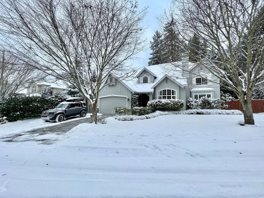 A suburban house with a snow-covered roof and yard, surrounded by bare trees.