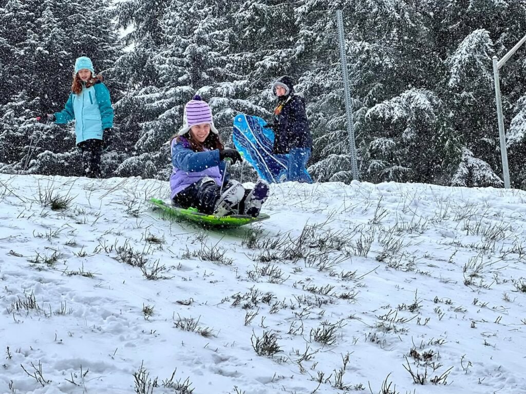 A person sledding down a snowy hill with others standing nearby in colorful winter clothing.